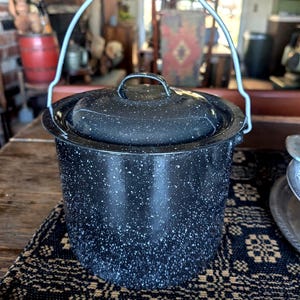 May include: A black enamel pot with a lid and a wire handle. The pot has a speckled white pattern and is sitting on a dark patterned mat. The pot is likely used for cooking.