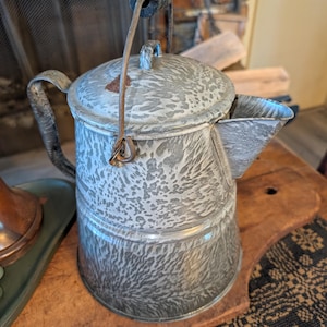 May include: Vintage speckled enamelware coffee pot with a black handle and spout. The pot has a gray and white pattern and a hinged lid. The handle is attached to the pot with a metal wire. The pot is sitting on a wooden surface.