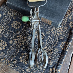May include: Vintage hand-crank egg beater with a wooden handle and a green knob. The beater is made of metal and has a classic design. It is resting on a patterned black and tan textile, with a dark book in the background.