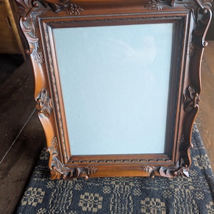 May include: A rectangular picture frame with a dark brown finish and ornate, carved detailing along the edges. The frame features a smooth, light-colored matting inside, providing a neutral border for a photograph or artwork display.
