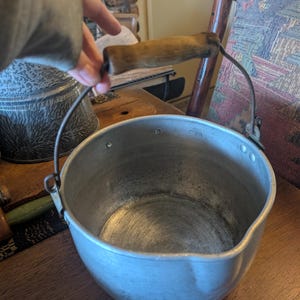 May include: A vintage aluminum cooking pot with a wooden handle and a metal wire handle. The pot has a rounded shape and a slightly flared rim. The interior shows signs of use. The handle is made of light brown wood.