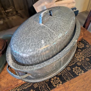 May include: Oval-shaped, gray enamelware casserole dish with a lid. The dish has a speckled pattern and a metal handle on the lid. The casserole dish is sitting on a wooden table with a patterned runner.