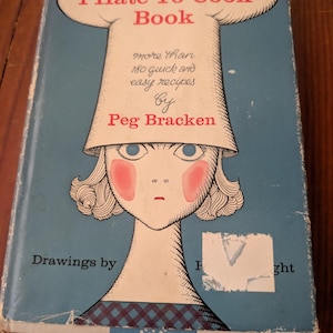 May include: A hardcover book titled The I Hate To Cook Book by Peg Bracken with a blue cover featuring a stylized illustration of a person wearing a large white chef hat, pink cheeks, and a blue checkered collar, with visible edge wear.