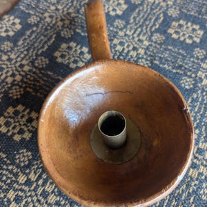 May include: A handcrafted wooden candle holder with a small handle. The candle holder is a warm brown color with a brass candle cup in the center. The candle holder is sitting on a blue and brown patterned surface.