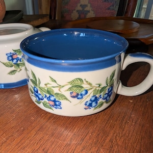May include: A white ceramic mug with a blue interior and handle. The mug features a floral design of blue berries and green leaves. The rim of the mug is also blue. Another similar mug is partially visible in the background.