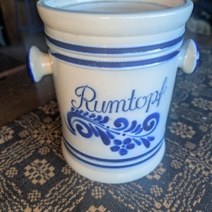May include: A white ceramic Rumtopf pot with blue stripes and handles. The pot features the word "Rumtopf" in blue script with floral accents. The pot is cylindrical with a slightly flared rim.