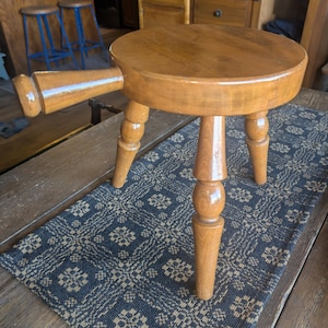 May include: A small, round wooden stool with a handle. The stool has three legs and a circular seat. The wood is a warm, light brown color. The handle extends from the side of the seat. The stool is on a patterned rug.