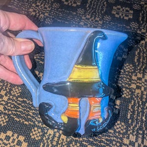 May include: A light blue ceramic mug featuring a raised, three-dimensional design of a witch hat in black and yellow, positioned above a pair of orange and yellow striped stockings ending in black curled-toe boots, all set against the blue mug body.