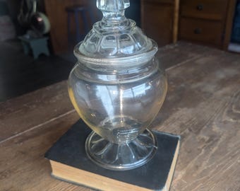 May include: A clear glass apothecary jar with a pointed finial lid. The jar is set on top of a dark book with yellowed pages. The jar has a round base and a bulbous body.