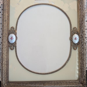 May include: An antique, square picture frame with a filigree metal border. The frame has an oval opening in the center and two decorative oval accents with floral designs. The frame is a vintage piece.