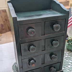 May include: A dark green wooden cabinet with eight small drawers and round knobs. The cabinet has a decorative top with a hole for hanging. The piece is sitting on a green lace doily.