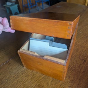 May include: A rectangular wooden index card box with finger joint corners and a hinged lid. The box is finished in a medium brown stain and contains a single light-colored, lined index card divider with a tab visible inside the open compartment.