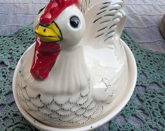 May include: A white ceramic hen-shaped dish with a red comb and wattle, and a yellow beak. The hen has black markings and is sitting on a matching white bowl. The dish is likely used for serving or storing food.