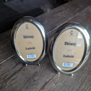 May include: Two oval-shaped brass-colored picture frames with metallic finishes and small feet. Each frame contains a beige insert with the text Columbia, The Brass Look, 4x6 inches, 10x15 cm, and Laiton, along with a barcode and product information.