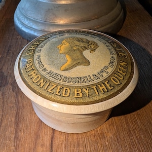 May include: A round, white, ceramic container with a gold and yellow lid. The lid features a woman's profile and the text "CHERRY TOOTH PASTE FOR BEAUTIFYING AND PRESERVING THE TEETH & GUMS LONDON PREPARED BY JOHN COSNELL & CO LTD PATRONIZED BY THE QUEEN".
