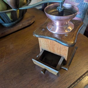 May include: A vintage manual coffee grinder with a wooden base and drawer. The grinder features a copper bowl and a metal crank handle. The drawer is open, revealing the interior. The grinder sits on a wooden surface.