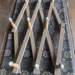 May include: A wooden accordion-style coat rack with multiple pegs. The rack is made of light brown wood with a ribbed design and is mounted on a dark patterned textile. The pegs have white tips.