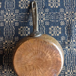 May include: A round copper pan featuring a hammered texture on the base and a long, straight metal handle with a hole at the top. The pan has a metallic, reddish-brown finish and is attached to the handle with two visible rivets at the base.