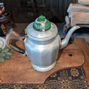 May include: A silver aluminum coffee pot with a green glass lid. The pot has a wooden handle and is sitting on a wooden board.