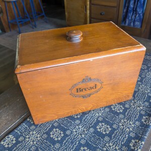 May include: A rectangular wooden bread box with a medium brown finish and a matching lid featuring a small round knob. The front panel displays the word Bread printed in a black decorative script within an ornate oval frame design.