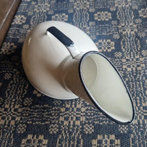 May include: Cream-colored enamelware pitcher with a black handle and rim. The pitcher has a rounded body and a spout. The background is a blue and tan patterned textile.
