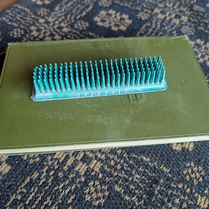 May include: A teal-colored floral frog, labeled "STEMGRIP", rests on a green book. The frog has many small, sharp pins for arranging flowers. The book is on a patterned surface.