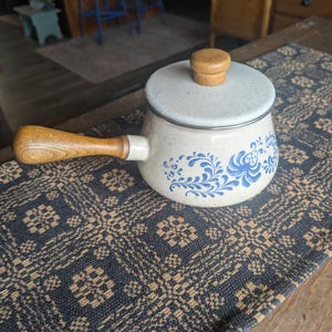 May include: A cream-colored speckled enamel saucepan with a matching lid, featuring a blue floral pattern around the base. The pot has a long, light brown wooden handle and a small wooden knob on the lid, providing a functional and classic design.