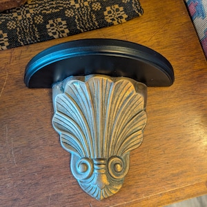 May include: A decorative wall shelf with a black, semi-circular top and a bronze-colored, seashell-shaped bracket. The bracket features intricate detailing and a scroll design. The shelf is mounted on a wooden surface.