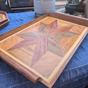May include: Wooden serving tray with a starburst design. The tray features a geometric star pattern in shades of brown and tan, framed by a lighter wood border. The tray is rectangular with a curved handle detail on one side.