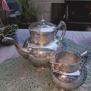 May include: A silver tea set, including a teapot with a lid and a creamer, sits on a green crocheted doily. The teapot and creamer have ornate floral patterns. The set is displayed outdoors.