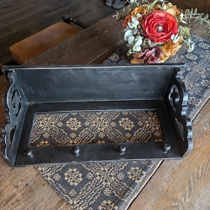 May include: A black wooden wall shelf with decorative cutouts and three small pegs. The shelf has a patterned fabric insert in shades of black and tan. A bouquet of flowers sits nearby on a patterned runner.
