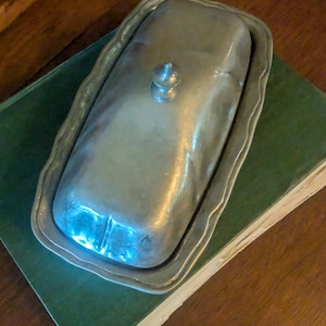 May include: A silver-colored butter dish with a lid and a decorative, scalloped edge. The dish sits on a green book, with a wooden surface in the background. The lid has a small, rounded handle.