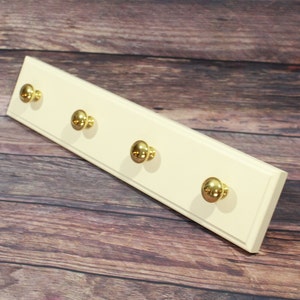 May include: A white wooden wall-mounted coat rack with four gold knob hooks.
