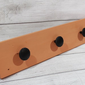 May include: A wooden coat rack with three black knobs. The rack is painted a light orange color.