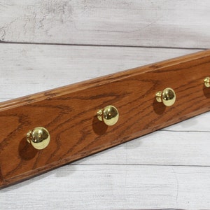 May include: A wooden wall-mounted coat rack with four brass-colored knobs. The rack is rectangular, made of stained wood, and has a beveled edge. The knobs are round and evenly spaced along the length of the rack.