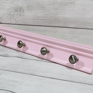 May include: A pink wooden wall-mounted coat rack with five silver metal hooks. The rack is approximately 18 inches long.