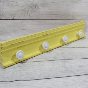 May include: A yellow wooden wall-mounted coat rack with four white ceramic knobs. The rack has a distressed finish and is mounted on a white wood background.