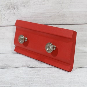 May include: A red wooden key holder with two silver knobs. The key holder is mounted on a white wood background.