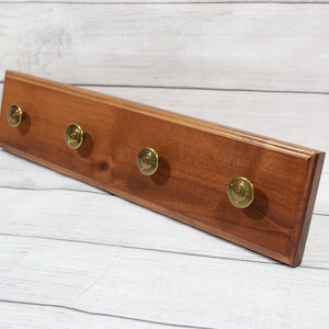 May include: A wooden wall-mounted key rack with four brass hooks. The rack is stained a dark brown color and has a slightly rounded edge.