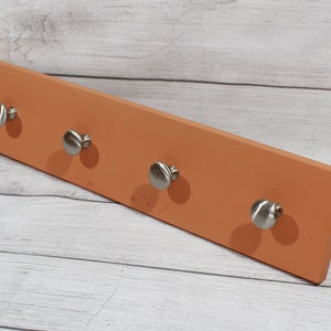 May include: A rectangular wooden wall-mounted coat rack with four silver metal hooks. The rack is painted a light orange color.