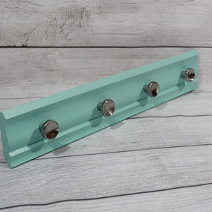 May include: A light blue wooden wall-mounted coat rack with five silver metal hooks. The rack is on a white wood background.