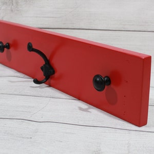 May include: Red wooden wall mounted coat rack with three hooks. Two hooks are black knobs and one is a black metal hook.