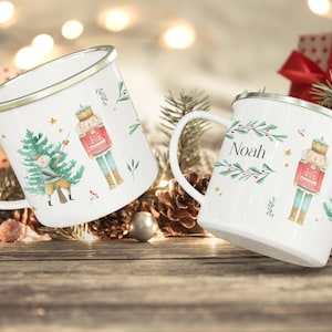 May include: Two white enamel mugs with a Christmas theme. One mug features a nutcracker and a Christmas tree with a star on top. The other mug has the name "Noah" and a nutcracker. Both mugs have a gold rim.