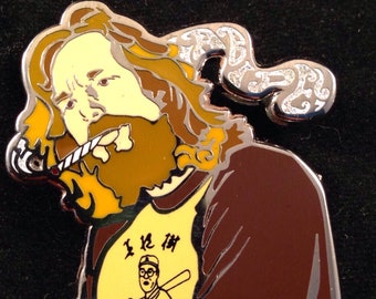 May include: A brown and yellow enamel pin featuring a cartoon character with long brown hair and a beard, smoking a cigarette. The character is wearing a brown shirt and a yellow t-shirt with a baseball player graphic and the text "I Love The".
