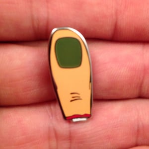 May include: A cartoon finger enamel pin with a green nail and a red band at the base.