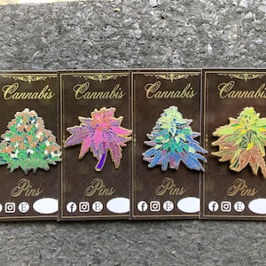 May include: Four colorful cannabis leaf pins on display cards. Each pin features a different color scheme: orange and green, pink and purple, blue and green, and yellow and red. The cards read "Cannabis" and "Pins".