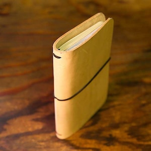 Può includere: Un diario in pelle color cuoio chiaro con chiusura elastica nera. Il diario è chiuso e ha il dorso arrotondato. Le pagine sono visibili in alto. Il diario è appoggiato su una superficie di legno.