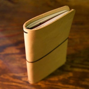 Può includere: Un diario in pelle beige con chiusura a fascia elastica nera. Le pagine sono visibili in alto. La pelle ha un aspetto naturale e leggermente strutturato. Il diario è in posizione verticale.