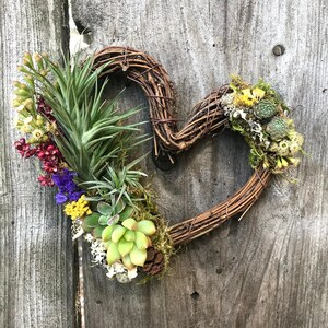 May include: A heart-shaped wreath made of brown twigs is decorated with green succulents, air plants, and dried flowers in shades of yellow, red, and purple.
