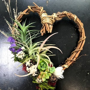 May include: A heart-shaped wreath made of brown twigs with a variety of green succulents, air plants, and dried flowers arranged inside.